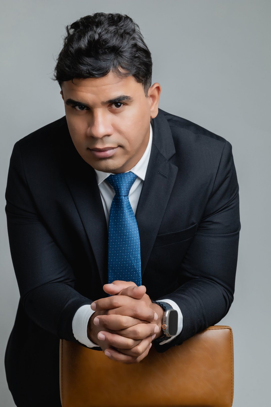 Dr. Alef Mascarenhas, de terno escuro e gravata azul, apoiado no encosto de uma cadeira